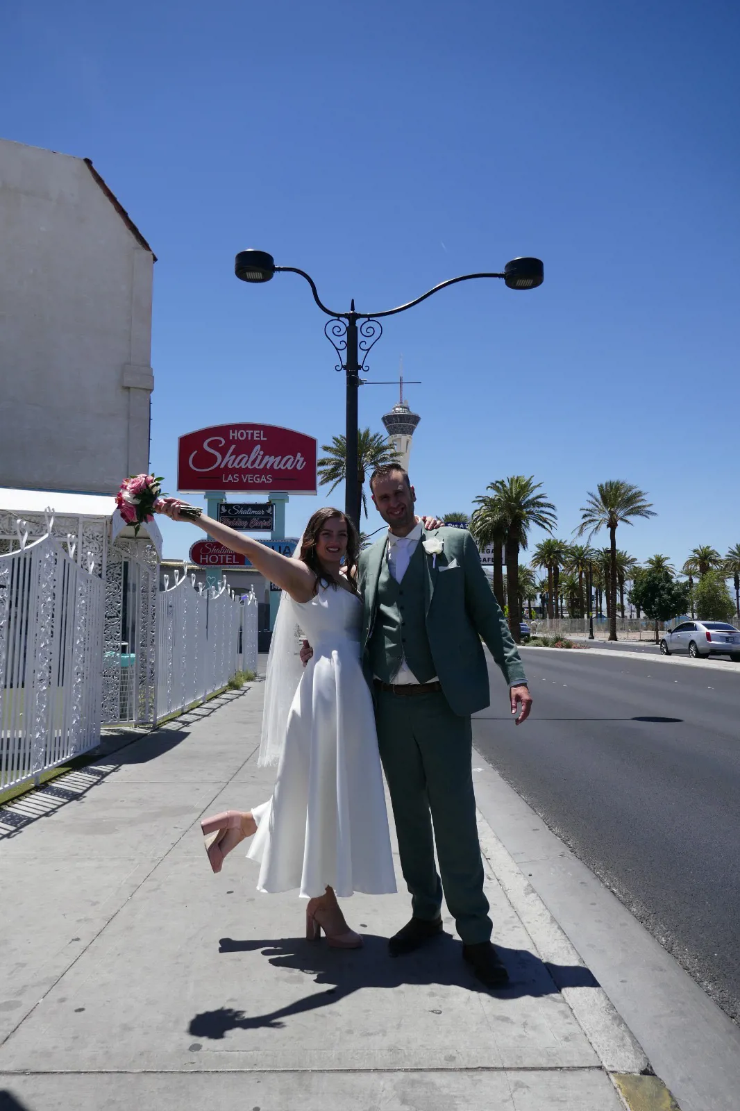 real bride lotte de lange – korte satijnen trouwjurk – bruid en bruidegom poseren stralend voor Hotel Shalimar in Las Vegas; bruid heft haar boeket omhoog terwijl ze speels één been optilt