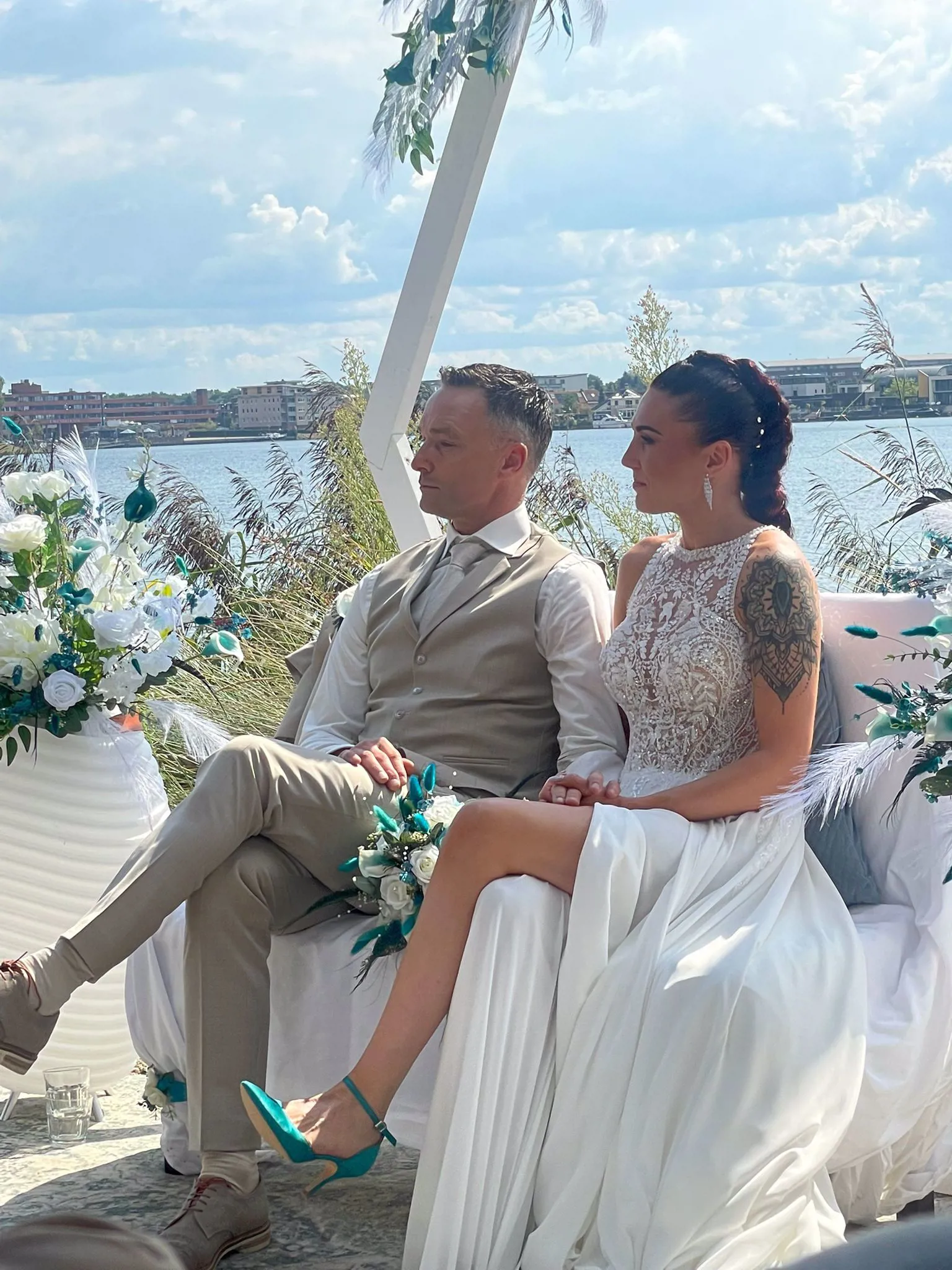 Bruid en bruidegom zitten samen tijdens hun ceremonie aan het water, bruid draagt een elegante trouwjurk van Bruidshuis Diana