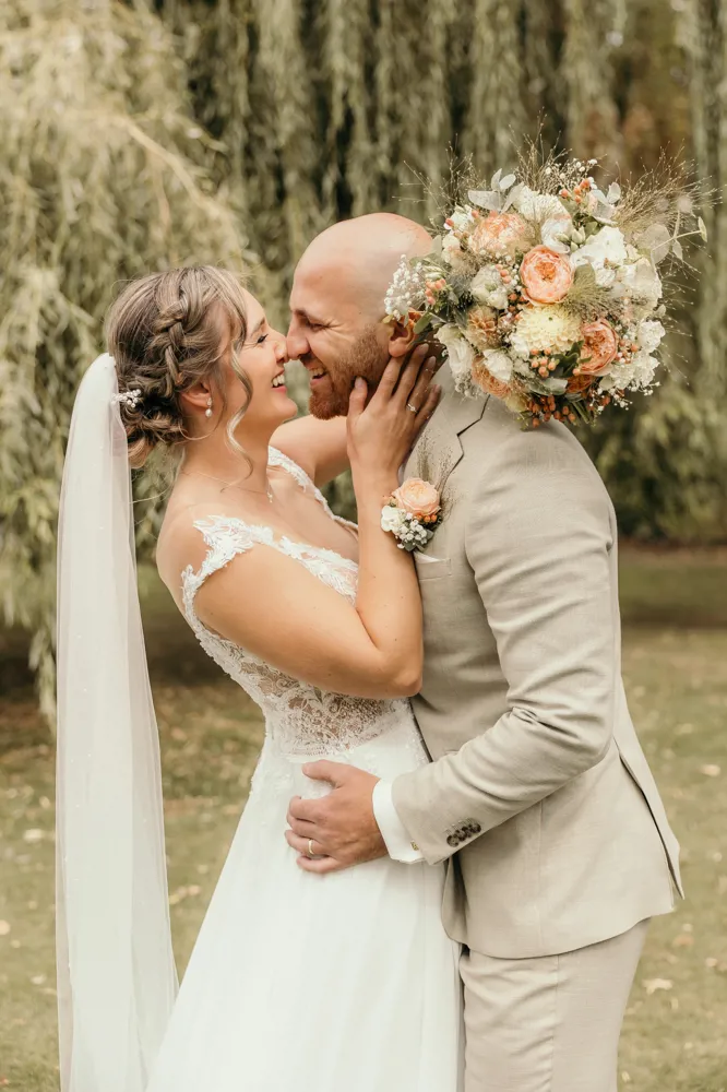 real-bride-review-babette-rosalle-bruidshuis-diana bruid Babette Rosalle lachend met haar partner onder treurwilg in romantische trouwjurk met kanten top en tule rok