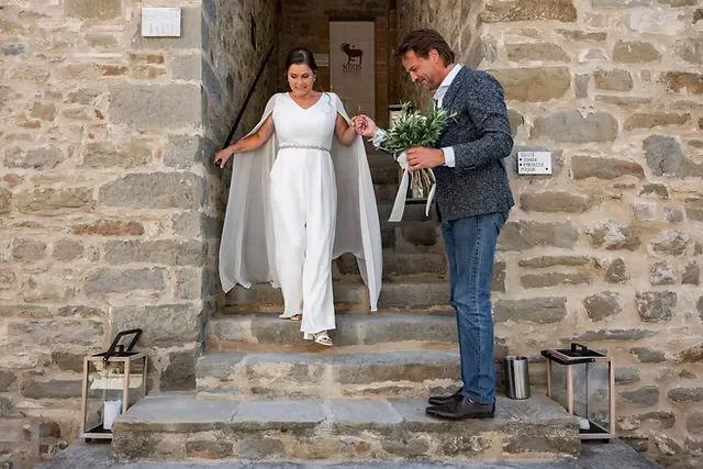real-bride-review-daphne-de-laat-bruidshuis-diana – bruid Daphne de Laat in een elegante ivoorkleurige jumpsuit met cape mouwen loopt hand in hand met haar bruidegom een stenen trap af tijdens hun bruiloft