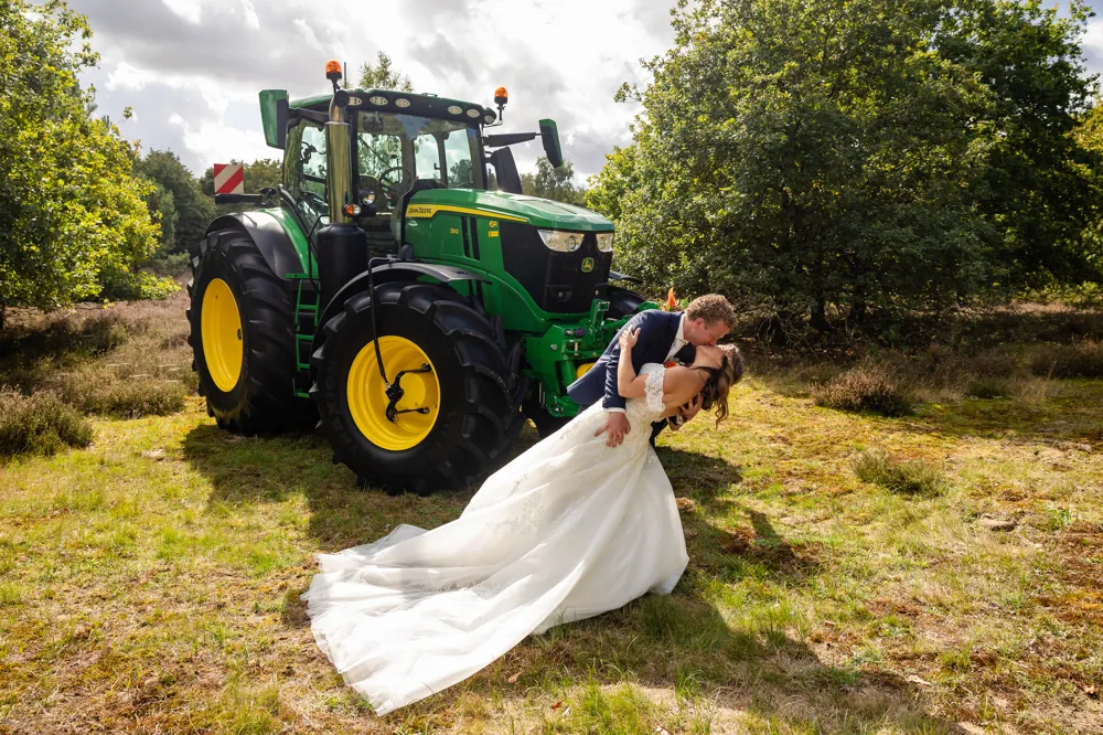 Bruid Birgit Arts wordt gekust door haar bruidegom bij een groene tractor in een landelijke setting, zij draagt een elegante trouwjurk van Bruidshuis Diana.