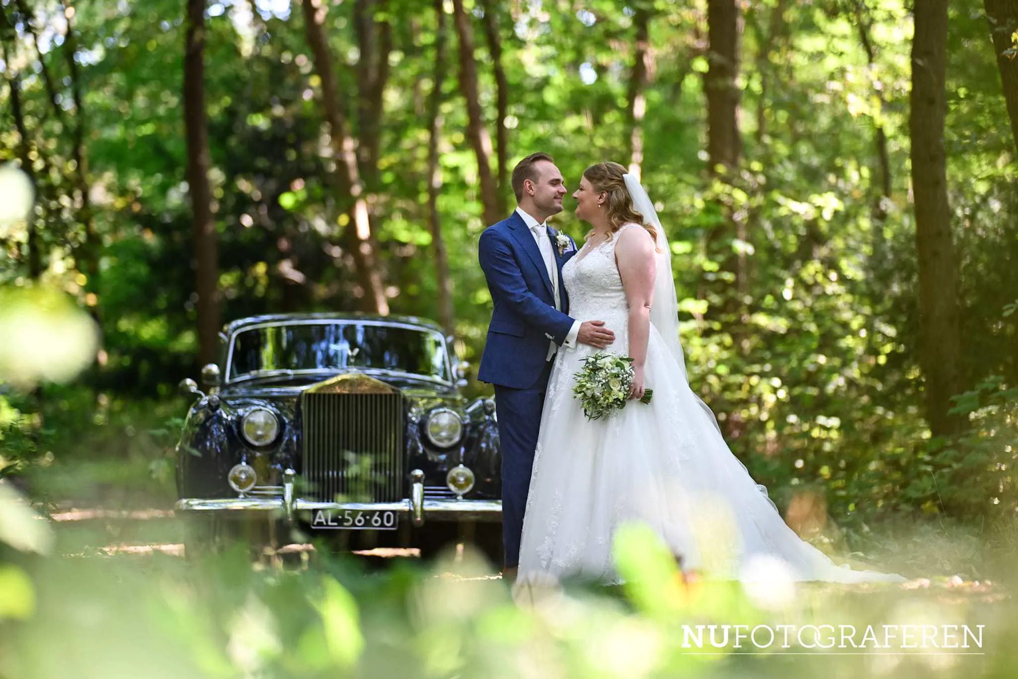Bruid Stefanie Hegelsom staat samen met haar bruidegom bij een klassieke zwarte auto in het bos en draagt een romantische trouwjurk van Bruidshuis Diana