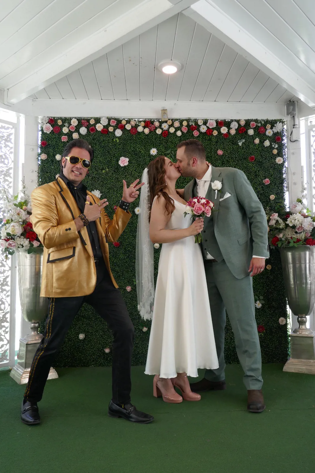 real bride lotte de lange – korte satijnen trouwjurk – bruid en bruidegom delen een kus bij een bloemenwand in A Little White Chapel, terwijl een Elvis-lookalike naast hen poseert