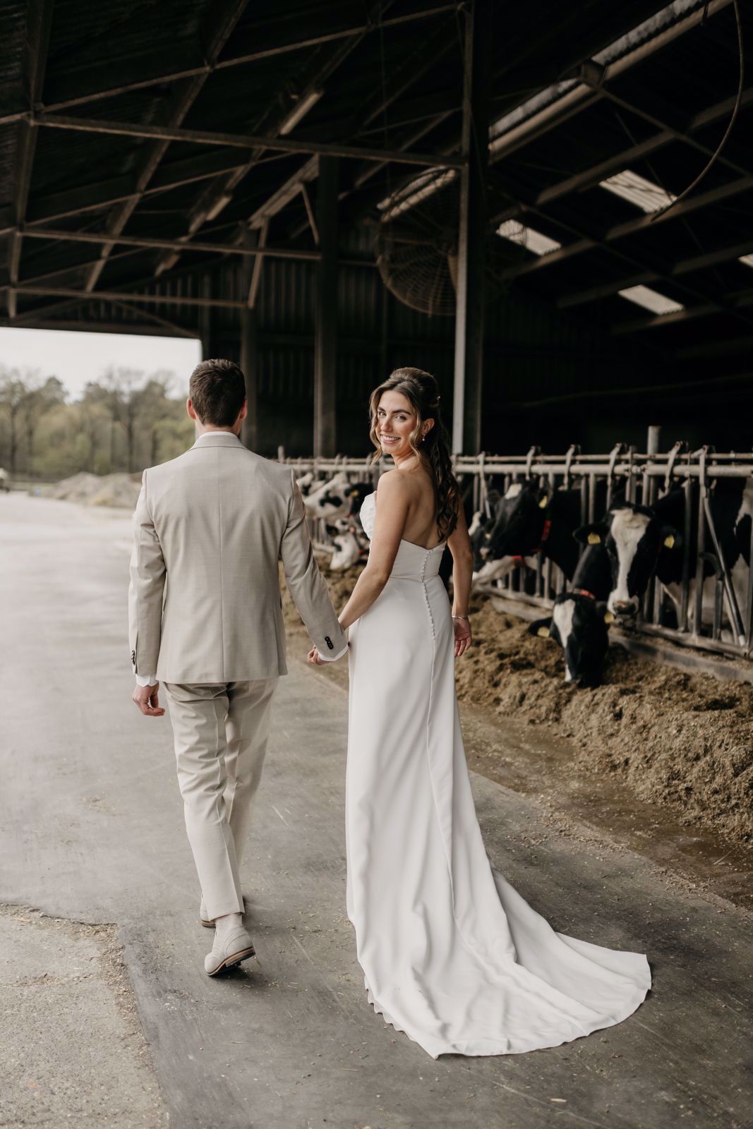 •	real bride Aafke Hoeben in een strakke, elegante trouwjurk met sleep, hand in hand met haar partner in een stal tussen de koeien – stoer en romantisch boerderijmoment 