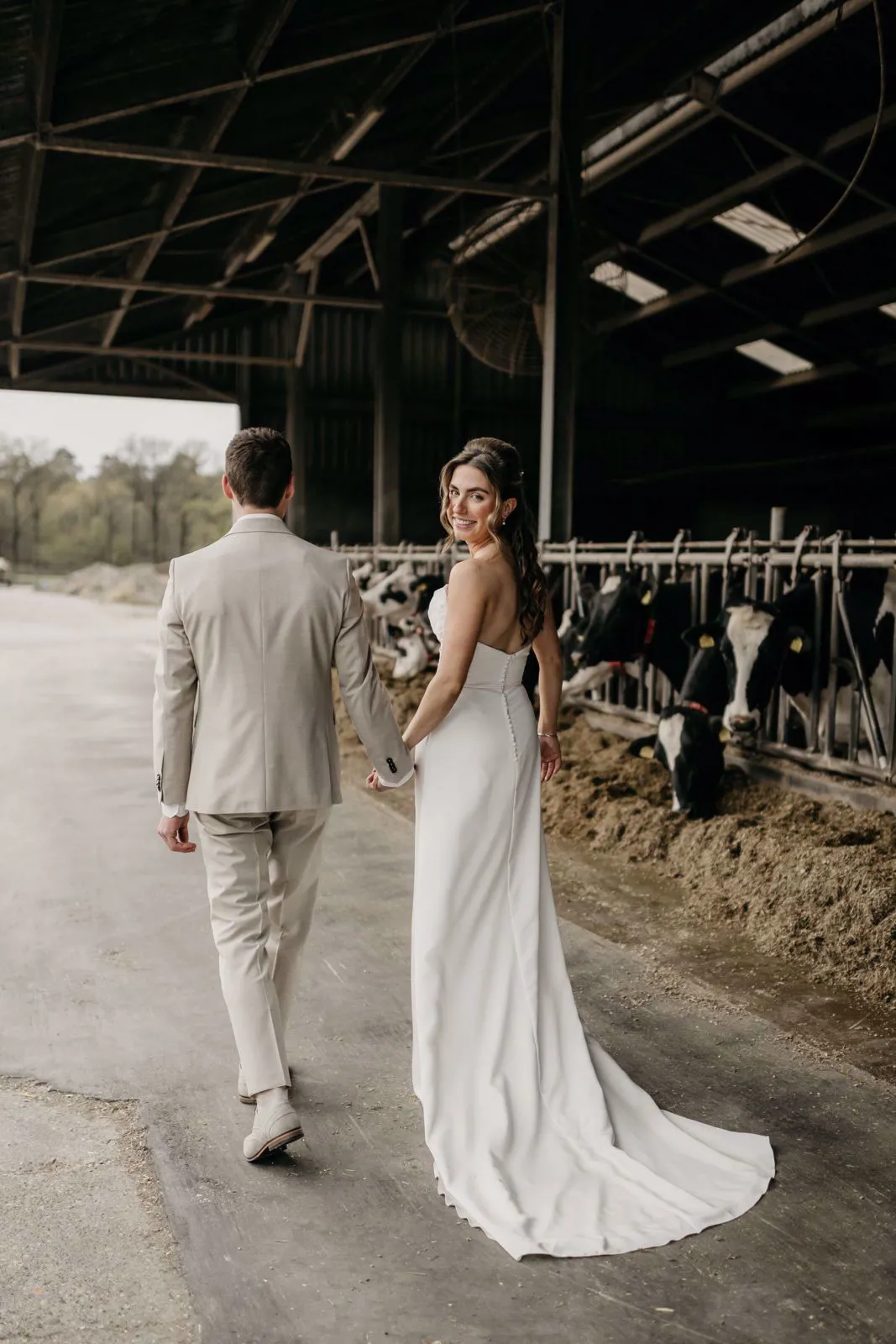 •	real bride Aafke Hoeben in een strakke, elegante trouwjurk met sleep, hand in hand met haar partner in een stal tussen de koeien – stoer en romantisch boerderijmoment 