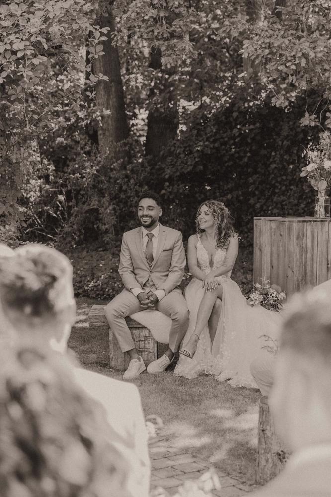 real bride stephanie van der linden – van der heijden – romantische A-lijn trouwjurk met split en kanten top – bruid en bruidegom zitten ontspannen tijdens de ceremonie onder de bomen (sepia/zwart-wit