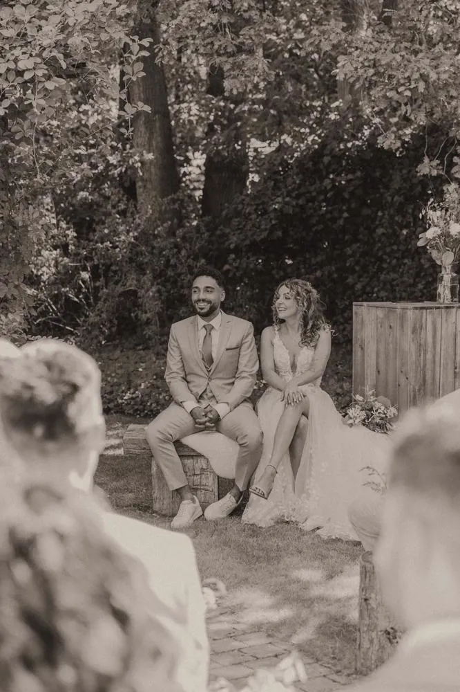 real bride stephanie van der linden – van der heijden – romantische A-lijn trouwjurk met split en kanten top – bruid en bruidegom zitten ontspannen tijdens de ceremonie onder de bomen (sepia/zwart-wit