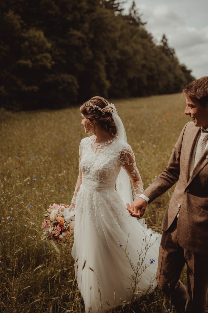 real-bride-review-geerdine-bruidshuis-diana – bruid Geerdine en haar bruidegom Wilbert staan samen in een zonnig landschap terwijl zij een elegante trouwjurk met kanten mouwen en lange sluier draagt en een kleurrijk bruidsboeket vasthoudt