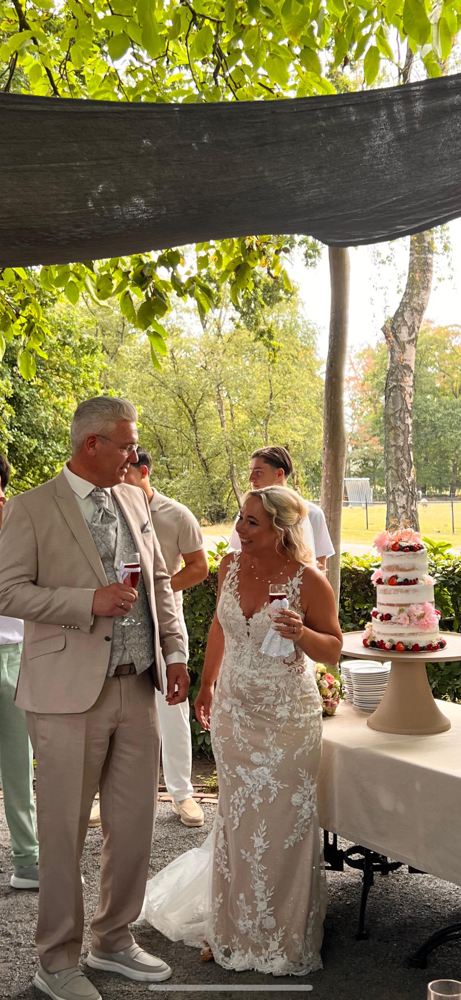 real bride cindy van der putten – elegante aansluitende trouwjurk met bloemenkant – bruid en bruidegom lachen naar elkaar tijdens het aansnijden van de taarten; romantische buitensetting met zomerse sfeer