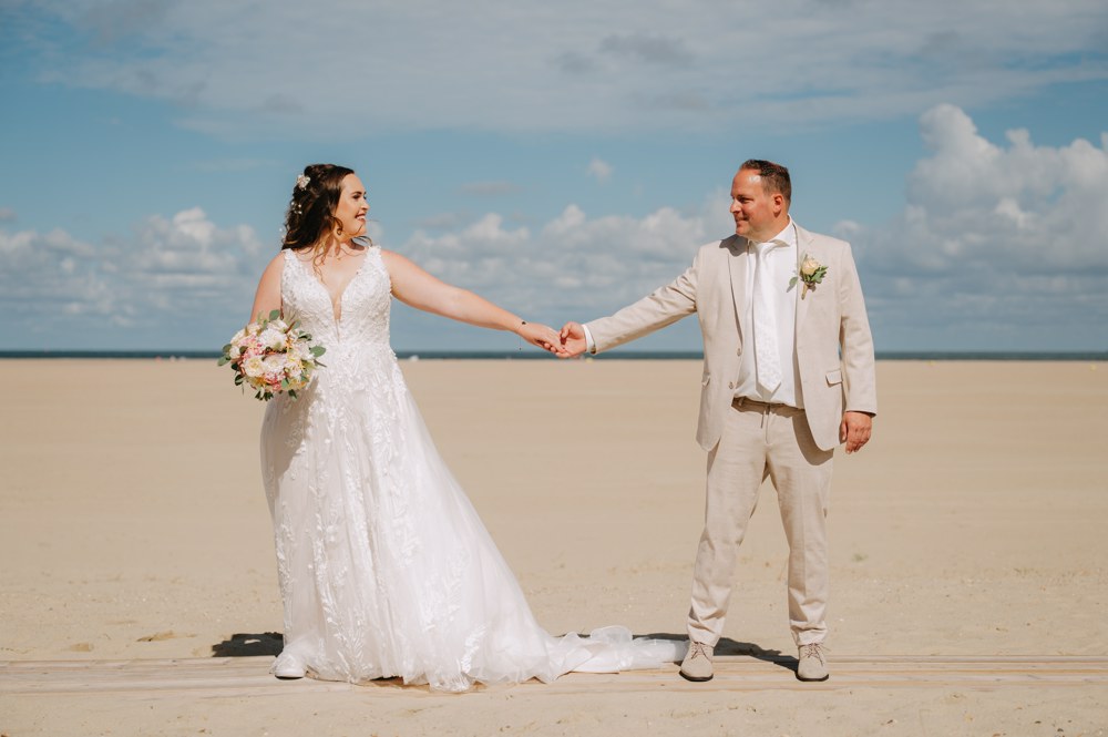 Bruid Lisette de Leeuw Egelmeers staat hand in hand met haar bruidegom op het strand, zij draagt een romantische kanten trouwjurk van Bruidshuis Diana.