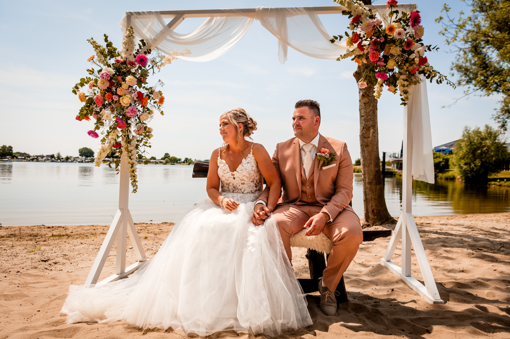 real-bride-review-marian-drost-bruidshuis-diana bruid en bruidegom onder sierlijke prieel omringd door strand en water op zonnige trouwdag