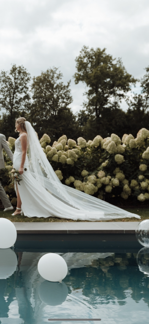 real bride daphne – fit & flare trouwjurk – bruid loopt buiten langs het zwembad; lange sleep en kanten sluier waaien elegant achter haar