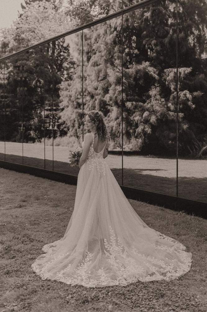 real bride stephanie van der linden – van der heijden – romantische A-lijn trouwjurk met lange sleep en bladapplicaties – bruid poseert sereen buiten voor een reflecterende muur (sepia/zwart-wit sfeer)