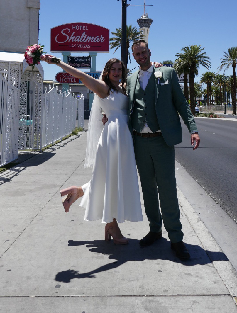 real bride lotte – korte satijnen trouwjurk – bruid en bruidegom poseren stralend voor Hotel Shalimar in Las Vegas; bruid heft haar boeket omhoog terwijl ze speels één been optilt