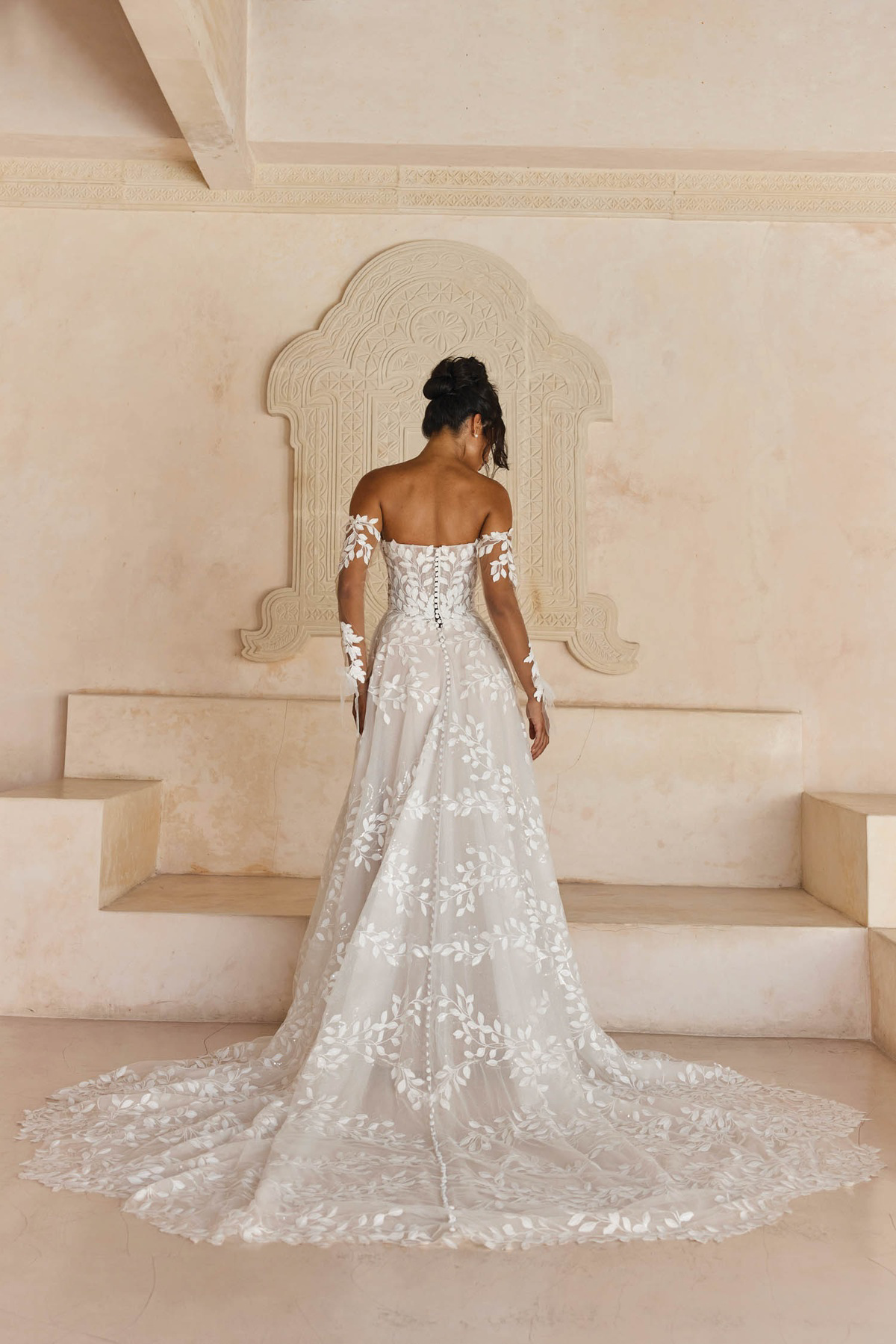 A-lijn trouwjurk Laiya van Madi Lane met bladmotief kant. Achteraanzicht met off-shoulder mouwen en een lange sleep.