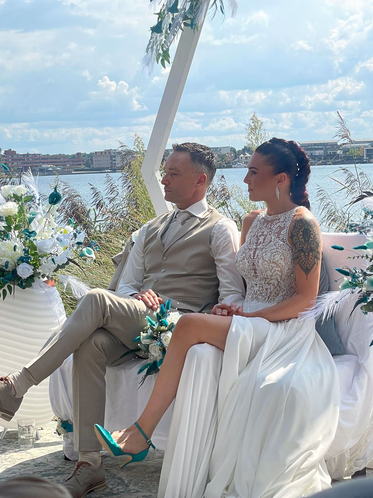 Bruid en bruidegom zitten samen tijdens hun ceremonie aan het water, bruid draagt een elegante trouwjurk van Bruidshuis Diana