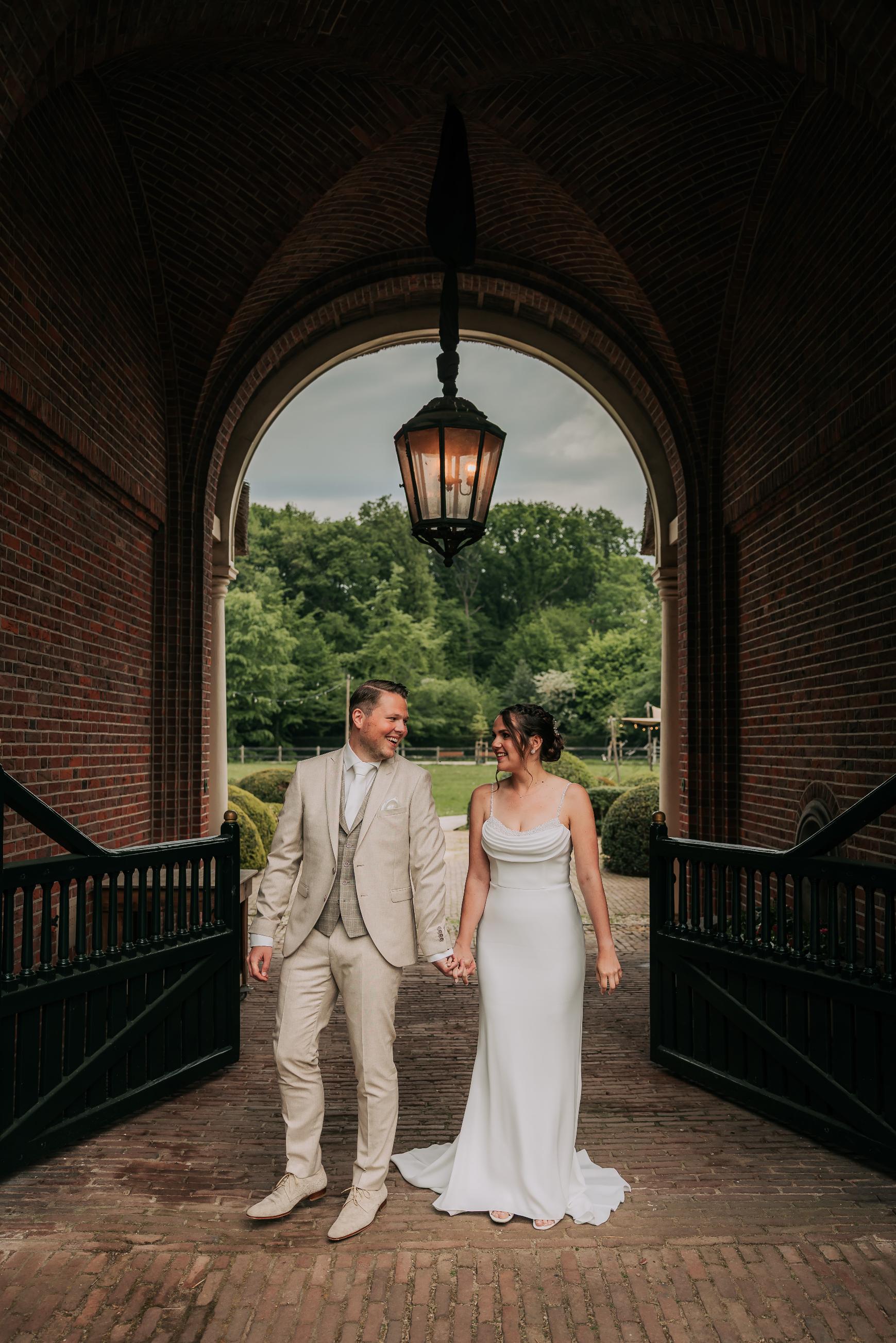 real-bride-review-emma-hesen-lommen-bruidshuis-diana – bruid Emma en bruidegom Stefan lopen hand in hand onder een poort terwijl zij een elegante minimalistische trouwjurk draagt