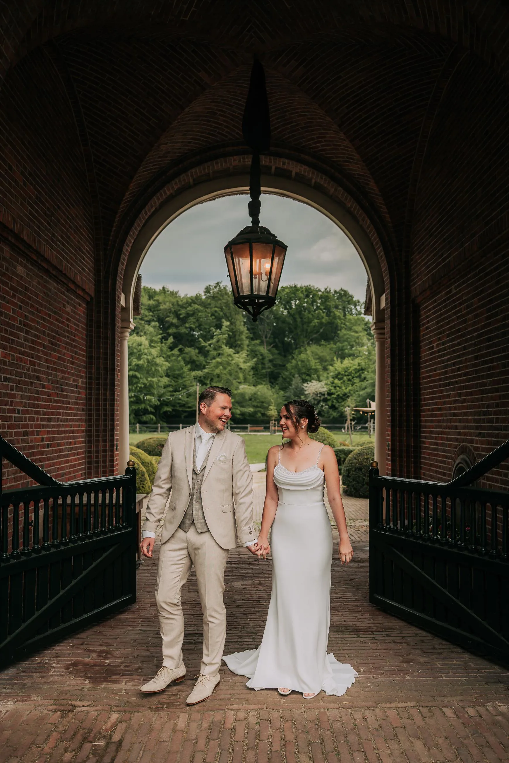 real-bride-review-emma-hesen-lommen-bruidshuis-diana – bruid Emma en bruidegom Stefan lopen hand in hand onder een poort terwijl zij een elegante minimalistische trouwjurk draagt