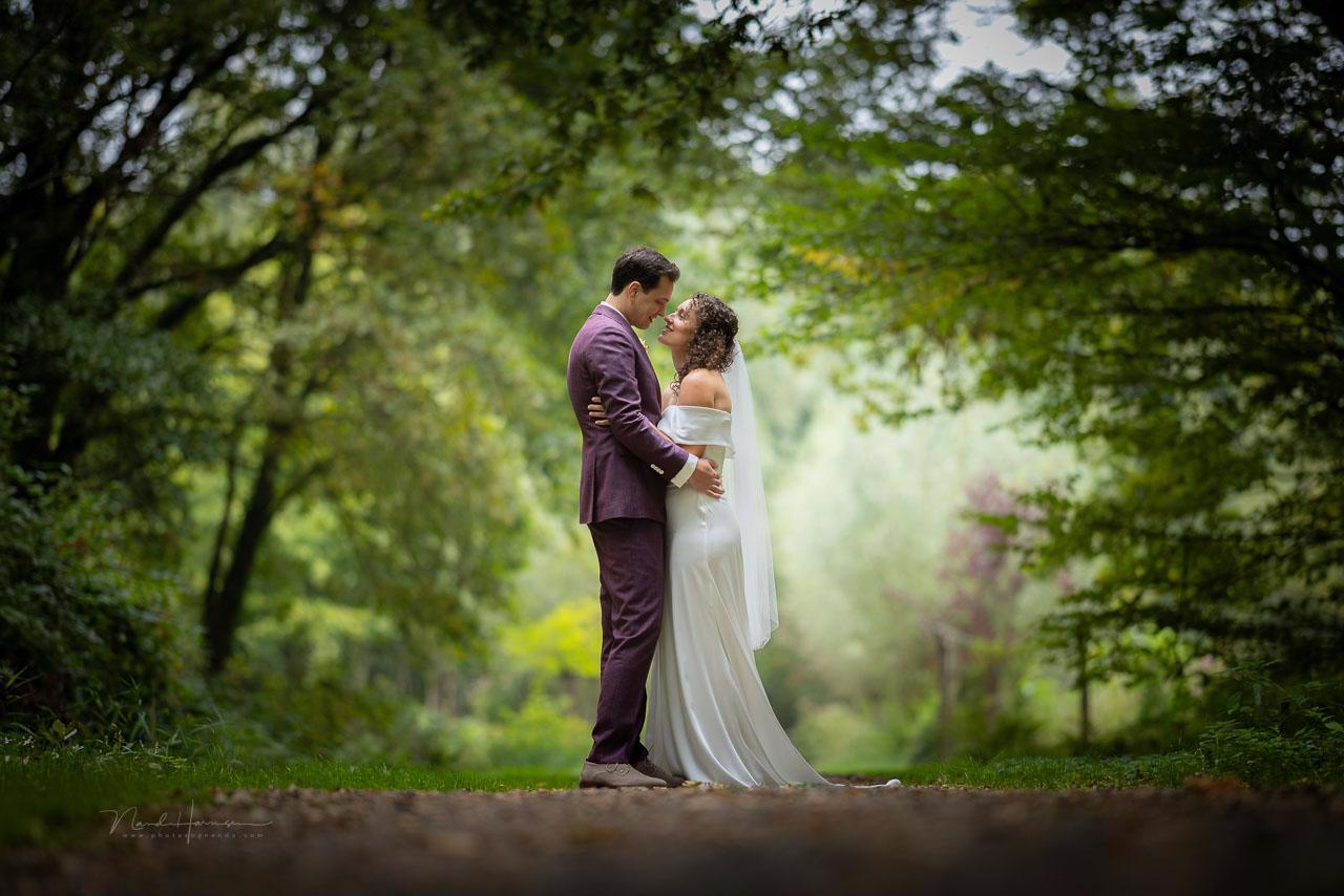 real bride loes martens – moderne fit & flare trouwjurk met off-shoulder draping – Loes en haar bruidegom staan liefdevol tegenover elkaar in een bosrijke omgeving; de sluier en vloeiende stof versterken het romantische moment