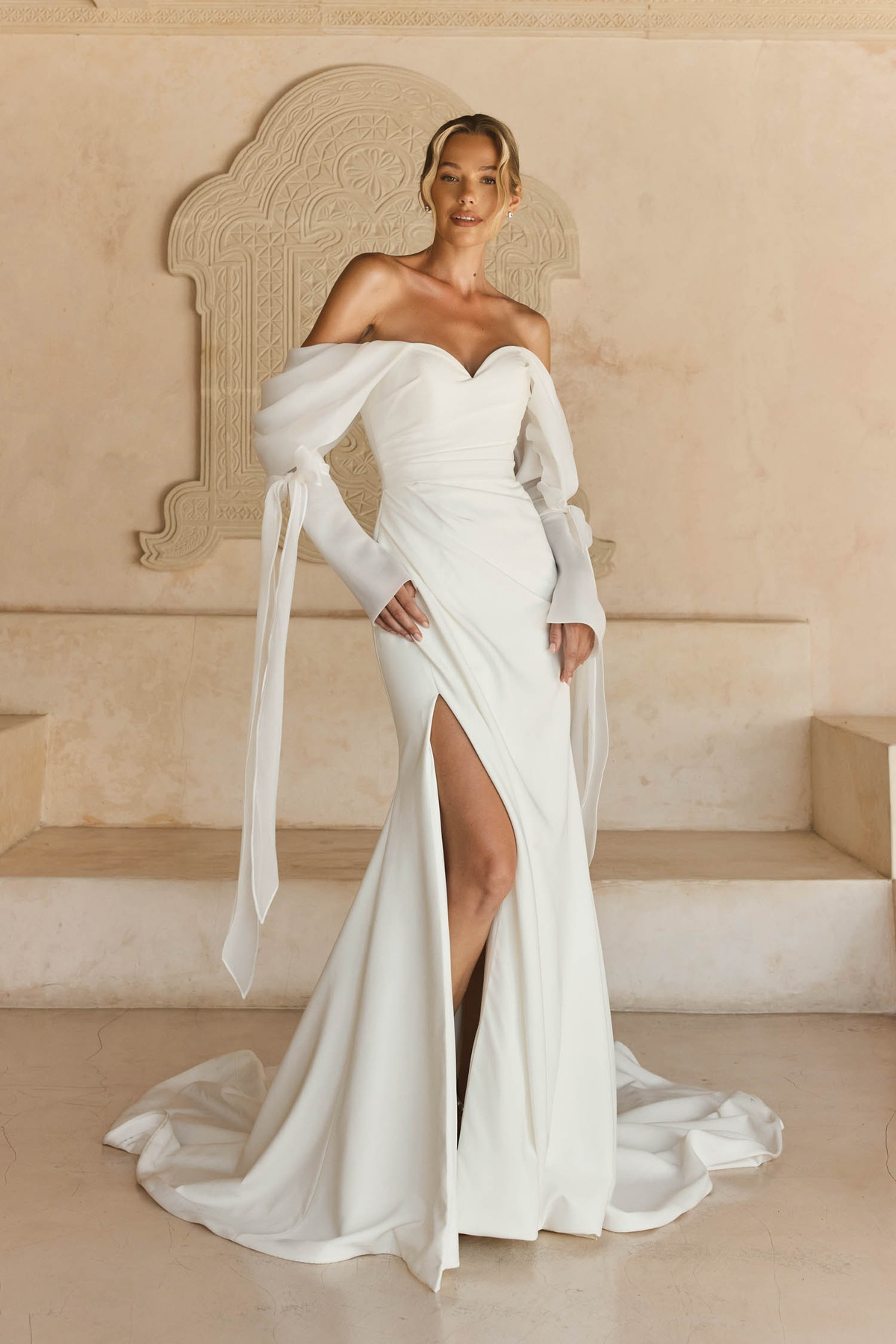 Off-shoulder trouwjurk Libbi van Madi Lane met split. Vooraanzicht van de aangesloten jurk met afneembare lange mouwen.