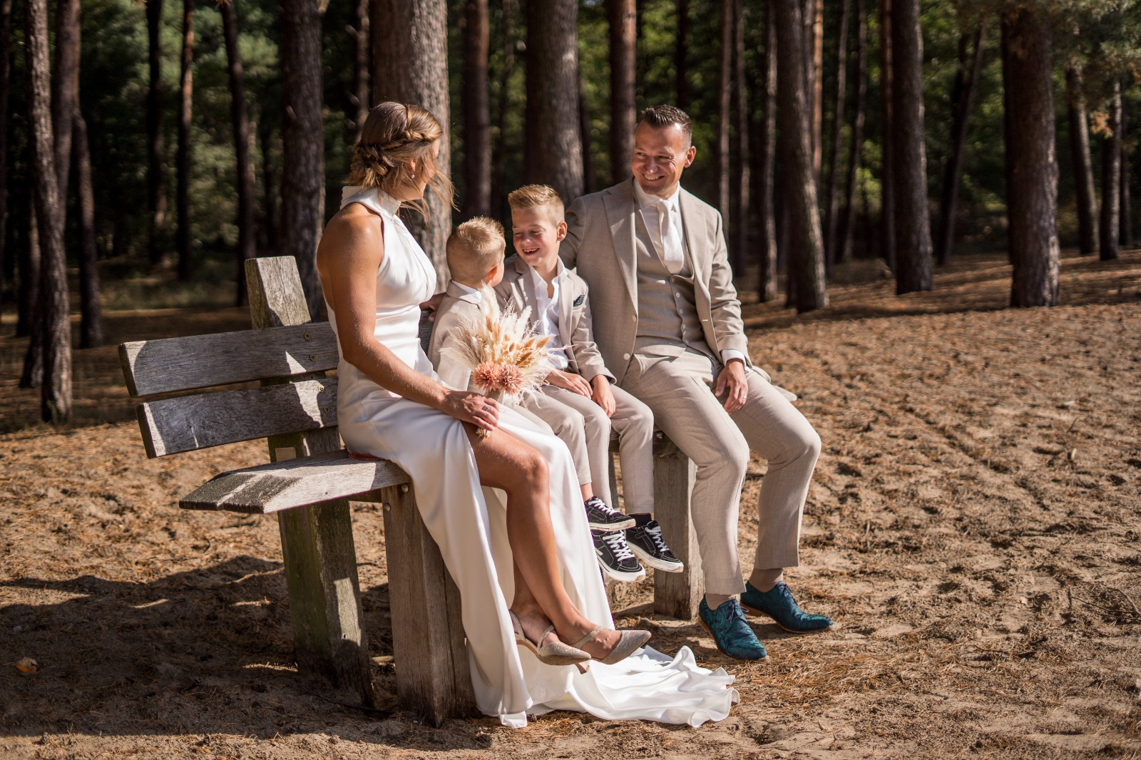 Bruid Mandy van Krey Lange zit samen met haar bruidegom en kinderen op een bankje in het bos, zij draagt een stijlvolle halter trouwjurk van Bruidshuis Diana.