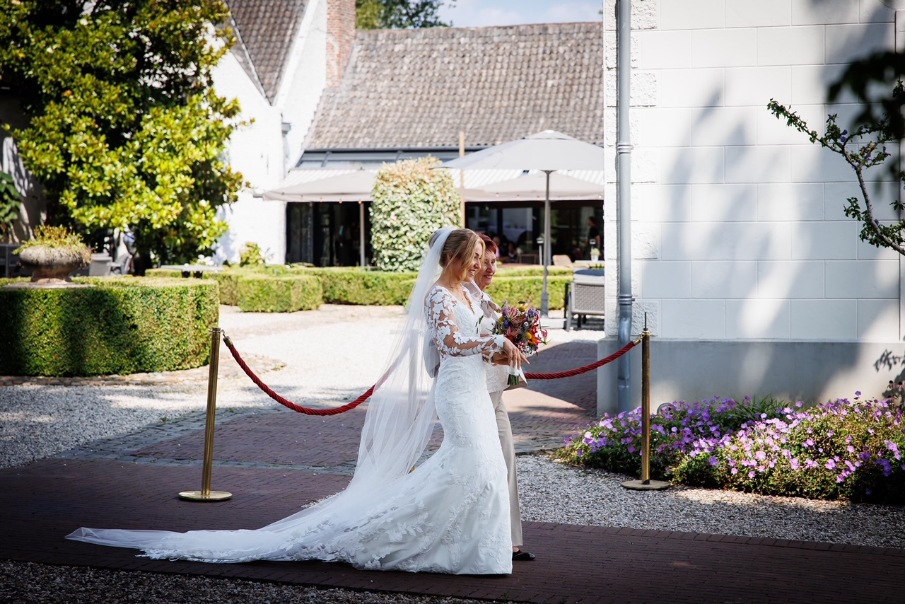 real-bride-review-fenna-van-gerven-bruidshuis-diana bruid Fenna van Gerven lopend hand naar ceremonie in kanten zeemeermin trouwjurk met open rug en lange sluier