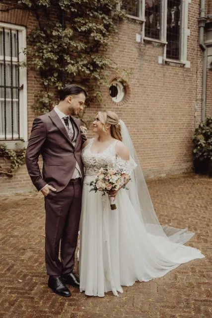 •	real bride Nirvana de Vrij in een romantische A-lijn trouwjurk met kanten top en tule rok, samen met haar partner in een bordeaux pak voor een historische bakstenen locatie 