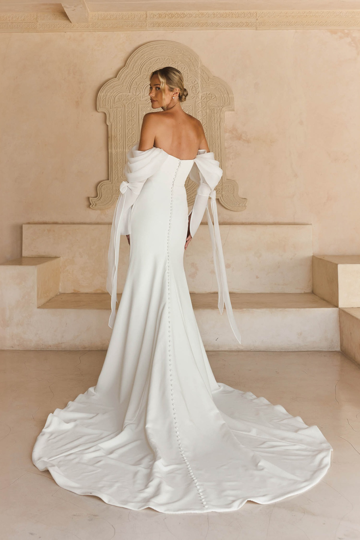 Off-shoulder trouwjurk Libbi van Madi Lane. Achteraanzicht van zeemeermin jurk met picpac knoopjes en strik mouwen.