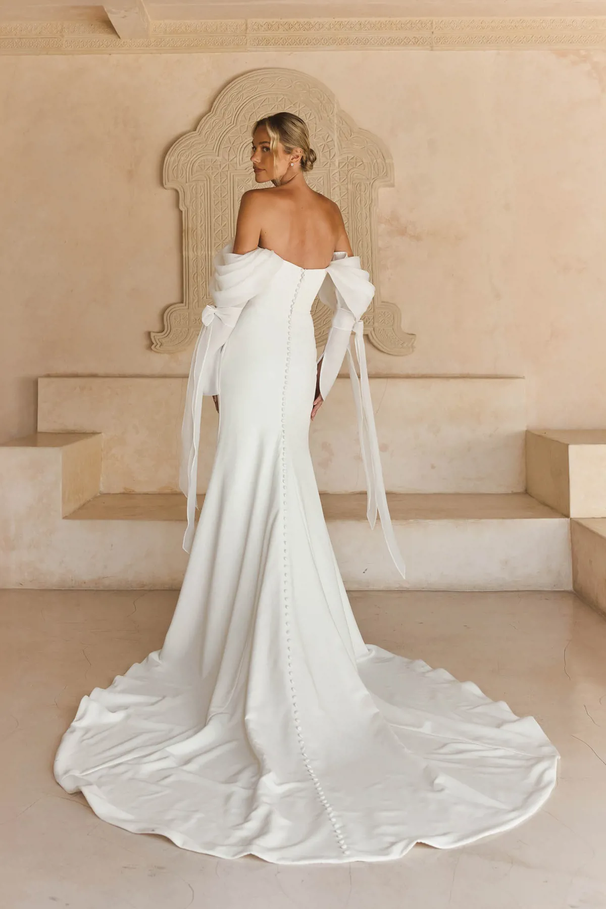 Off-shoulder trouwjurk Libbi van Madi Lane. Achteraanzicht van zeemeermin jurk met picpac knoopjes en strik mouwen.