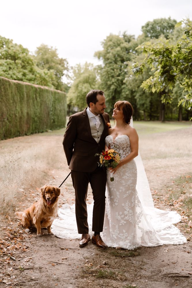 real-bride-review-amber-krikels-van-der-heijden-bruidshuis-diana – bruid Amber Krikels van der Heijden en haar bruidegom wandelen samen met hun hond over een pad terwijl zij een elegante strapless trouwjurk met bloemenapplicaties draagt