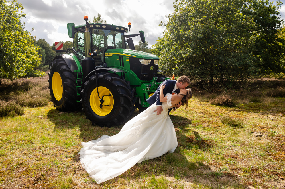 Bruid Birgit Arts wordt gekust door haar bruidegom bij een groene tractor in een landelijke setting, zij draagt een elegante trouwjurk van Bruidshuis Diana.