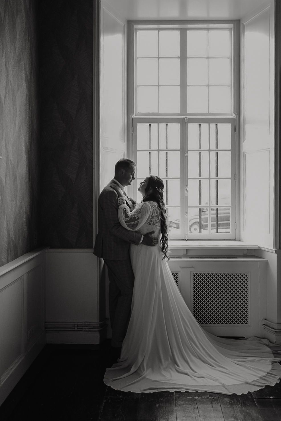 real-bride-review-hannah-bruidshuis-diana – bruid Hannah en haar bruidegom staan liefdevol tegenover elkaar bij een groot raam terwijl zij een elegante trouwjurk met kanten mouwen en lange rok draagt in een sfeervolle zwart-wit foto