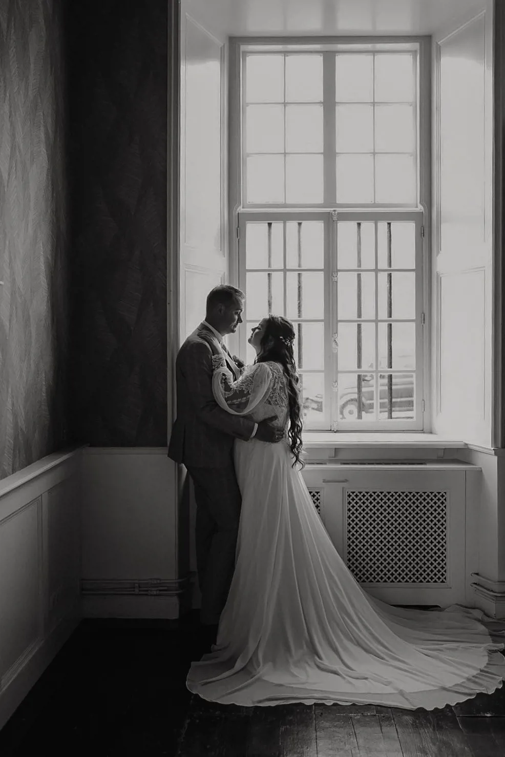 real-bride-review-hannah-bruidshuis-diana – bruid Hannah en haar bruidegom staan liefdevol tegenover elkaar bij een groot raam terwijl zij een elegante trouwjurk met kanten mouwen en lange rok draagt in een sfeervolle zwart-wit foto