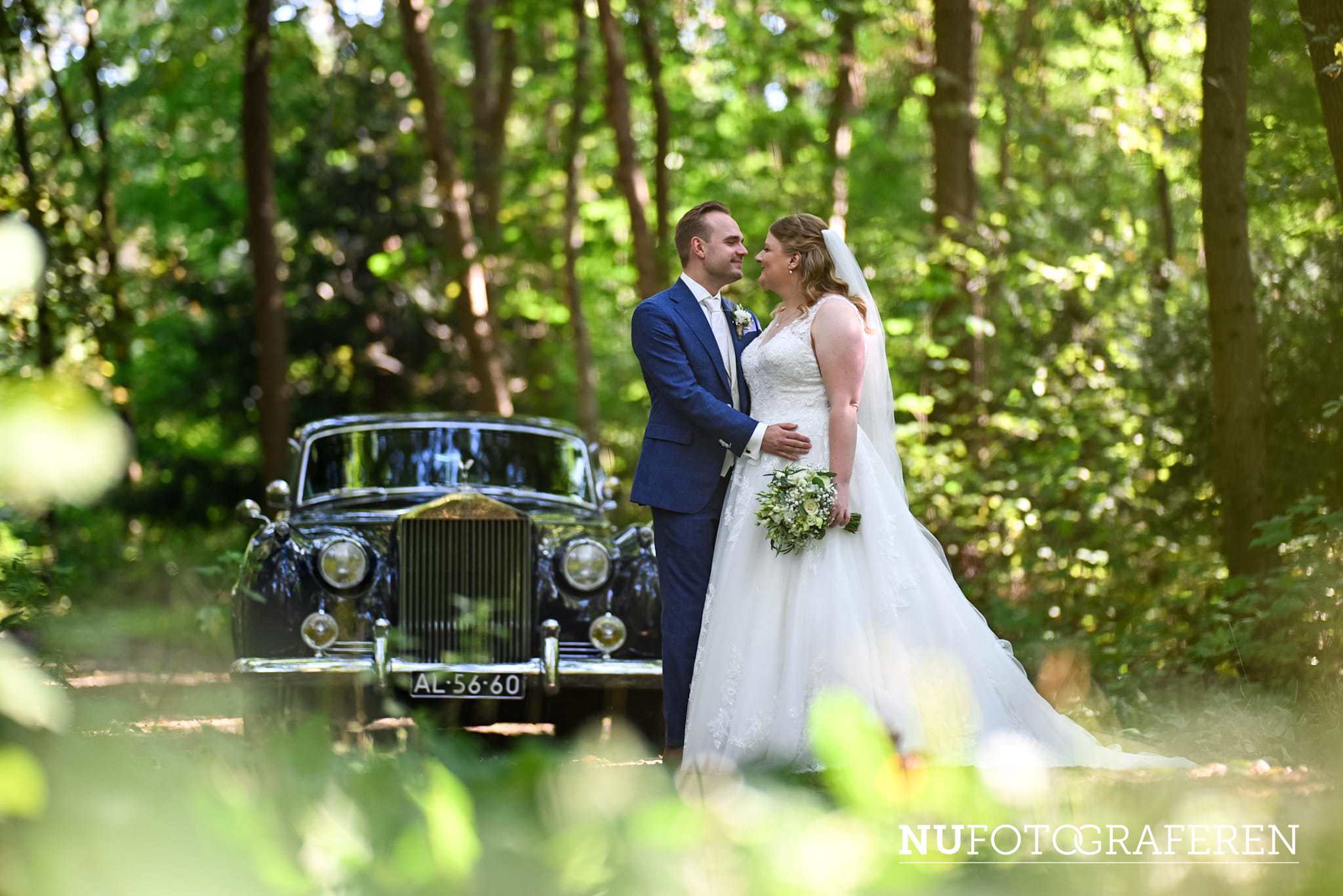 Bruid Stefanie Hegelsom staat samen met haar bruidegom bij een klassieke zwarte auto in het bos en draagt een romantische trouwjurk van Bruidshuis Diana