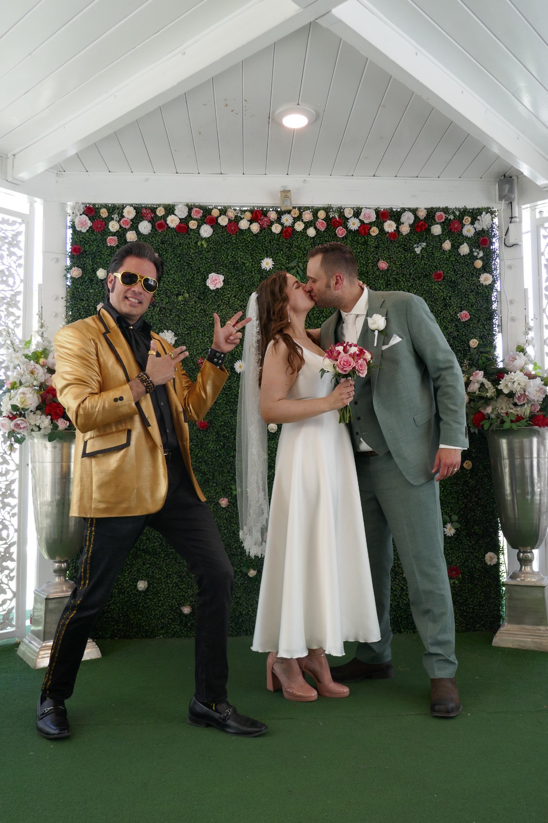 real bride lotte de lange – korte satijnen trouwjurk – bruid en bruidegom delen een kus bij een bloemenwand in A Little White Chapel, terwijl een Elvis-lookalike naast hen poseert