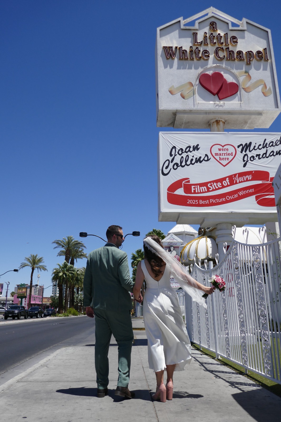 real bride lotte de lange – korte satijnen trouwjurk – bruid en bruidegom lopen hand in hand richting A Little White Chapel in Las Vegas; haar sluier waait achter haar terwijl ze haar boeket vasthoudt