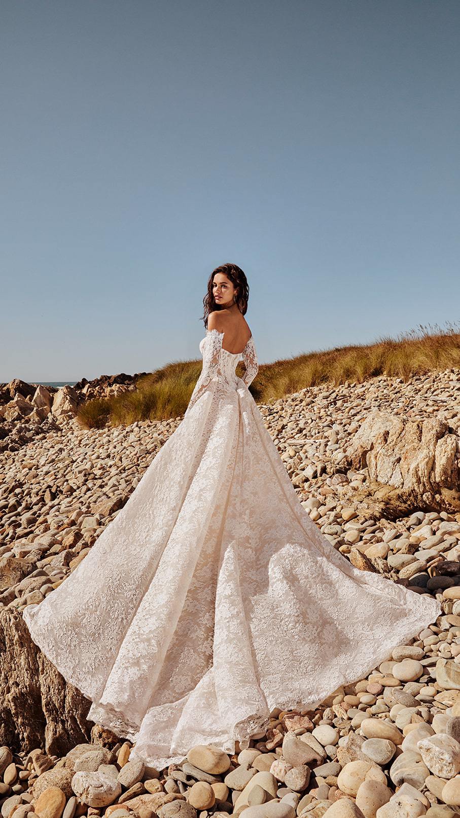 Rug van de Bridal Gala Akira jurk van Galia Lahav met uitlopende kanten rok, prachtige sleep en off-shoulder mouwen op een rotsachtig strand.